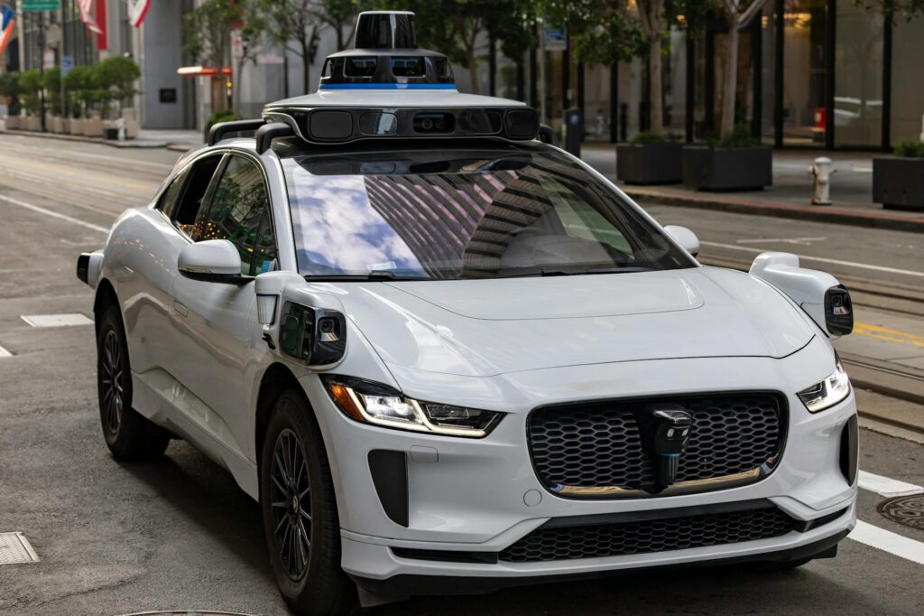 A white autonomous vehicle navigating a city street, reflecting urban architecture in daylight.